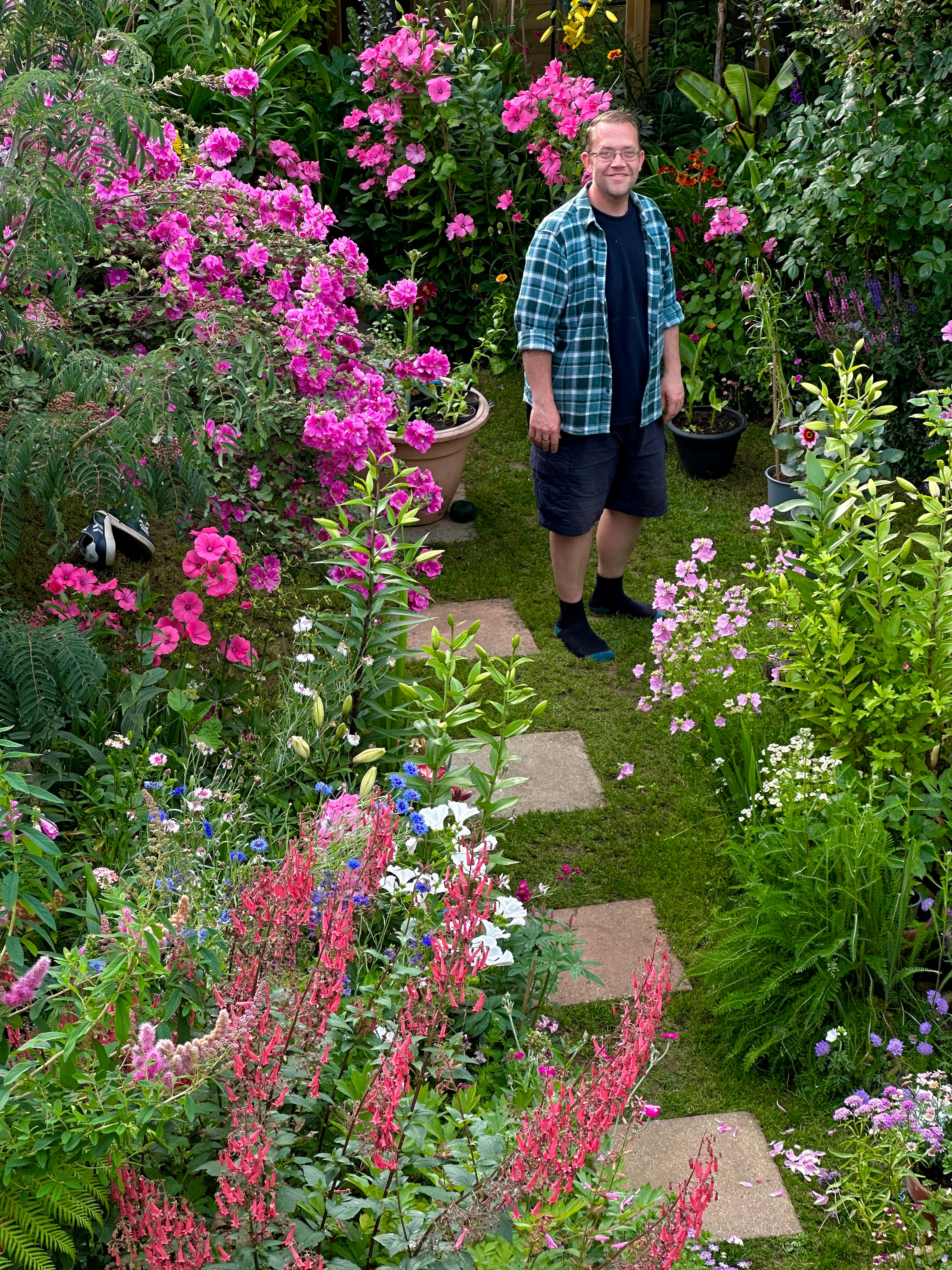 A man wearing a green and blue plaid flannel shirt, dark shorts, and socks stands on a stone path in a lush, vibrant garden filled with pink flowers and green foliage.